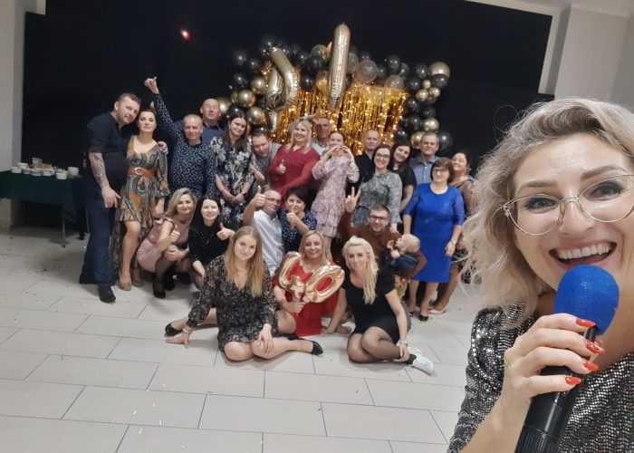 Duża grupa dorosłych świętuje, prawdopodobnie rocznicę, w sali ozdobionej czarno-złotymi balonami z cyframi "30". Na pierwszym planie uśmiechnięta kobieta (prawdopodobnie wodzirej lub animatorka) z blond włosami w okularach, trzymająca mikrofon, robi sobie selfie z całą grupą w tle.