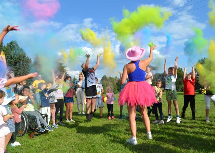 Duża grupa dorosłych i dzieci, w tym osoba na wózku inwalidzkim, rzuca w powietrze kolorowy proszek (prawdopodobnie holi) na trawniku w słoneczny dzień. W powietrzu unoszą się chmury różowego, żółtego, zielonego i niebieskiego proszku. Animatorka w różowym kowbojskim kapeluszu i różowej spódnicy tutu stoi tyłem do aparatu.