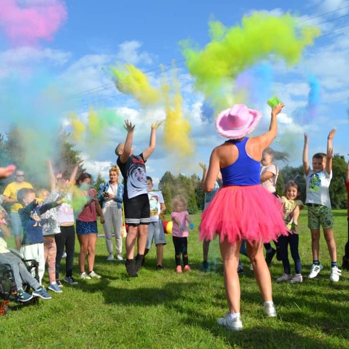Duża grupa dorosłych i dzieci, w tym osoba na wózku inwalidzkim, rzuca w powietrze kolorowy proszek (prawdopodobnie holi) na trawniku w słoneczny dzień. W powietrzu unoszą się chmury różowego, żółtego, zielonego i niebieskiego proszku. Animatorka w różowym kowbojskim kapeluszu i różowej spódnicy tutu stoi tyłem do aparatu.