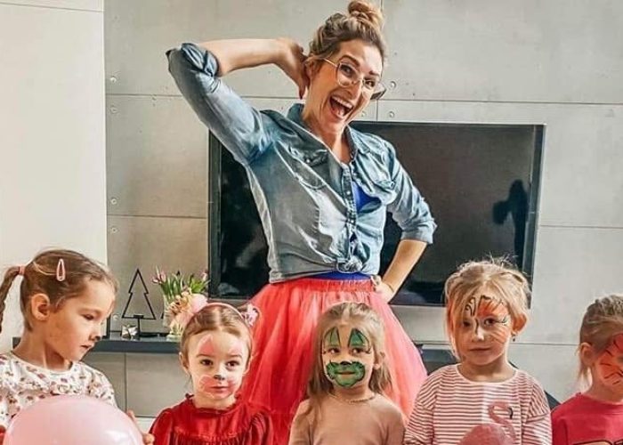 Kobieta w dżinsowej koszuli i różowej spódniczce tutu stoi, uśmiechając się szeroko i trzymając rękę za głową. Przed nią, od pasa w dół, siedzi czworo małych dzieci, z których dwoje ma pomalowane twarze (jedno na zielono, drugie w motywy zwierzęce). Scena rozgrywa się w nowoczesnym salonie.
