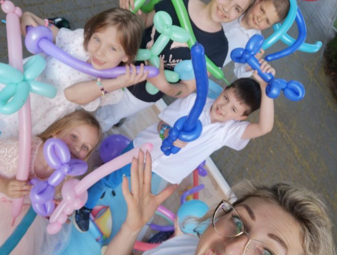 Zbiorowe selfie dorosłej kobiety i pięciorga dzieci, trzymających kolorowe balonowe figurki (miecze, kwiaty, pająka). Kobieta trzyma aparat i uśmiecha się szeroko, a dzieci są nad nią i wokół niej.
