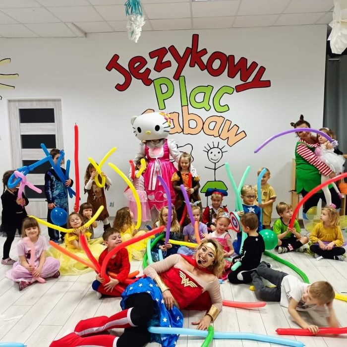 Duża grupa dzieci i dwoje dorosłych animatorów (jedna przebrana za Wonder Woman, druga w kostiumie Hello Kitty) siedzą lub leżą na podłodze w sali zabaw, otoczeni długimi, kolorowymi balonami. Na ścianie w tle widnieje kolorowy napis: "Językowy Plac Zabaw".