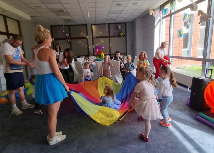 Zdjęcie z przyjęcia dla dzieci odbywającego się w przestronnej sali z dużymi oknami. Dwie animatorki, jedna w niebieskiej tiulowej spódnicy i druga w czerwonej koszulce, prowadzą zabawę z użyciem kolorowej chusty animacyjnej (Klanza). Wokół chusty bawią się dzieci, a w tle widać dekoracje z balonów i rozłożony sprzęt do zabaw.