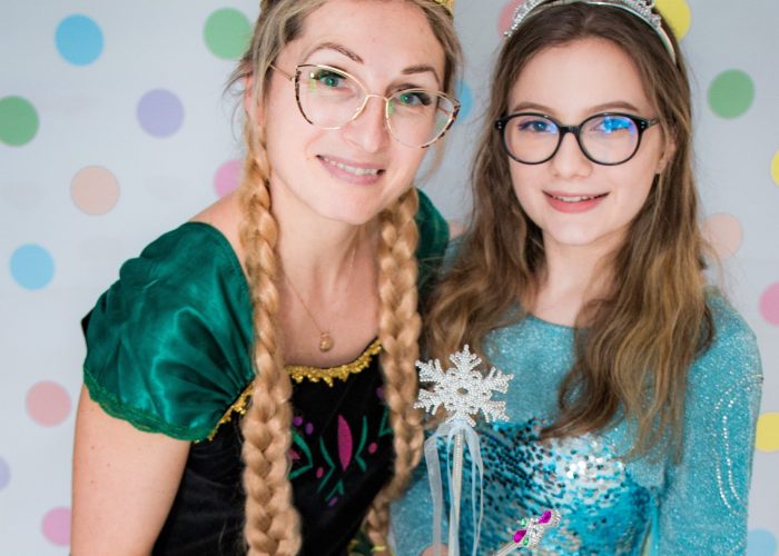 Kobieta i dziewczynka ubrane w stroje postaci z "Krainy Lodu" pozują przed ścianą w kolorowe kropki. Kobieta po lewej jest przebrana za Annę, w zielonej sukience i koronie, z zaplecionymi warkoczami. Dziewczynka po prawej jest przebrana za Elsę, w niebieskiej, błyszczącej sukience i srebrnej koronie, trzymając różdżkę w kształcie płatka śniegu. Obie mają okulary.