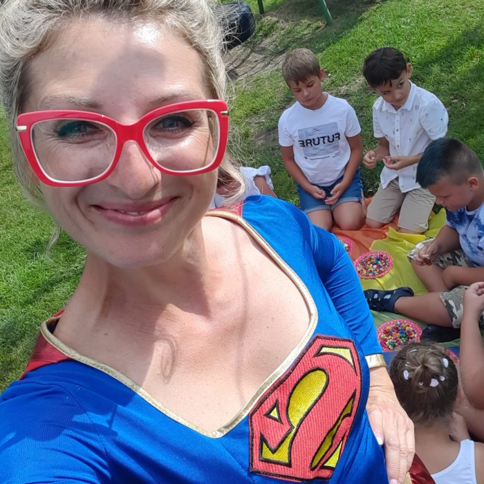 Selfie animatorki przebranej za Superwoman (w niebieskim stroju z symbolem "S" na piersi i czerwoną peleryną) w okularach w czerwonych oprawkach. Uśmiecha się do aparatu. W tle, na trawie, siedzi i bawi się grupa chłopców.